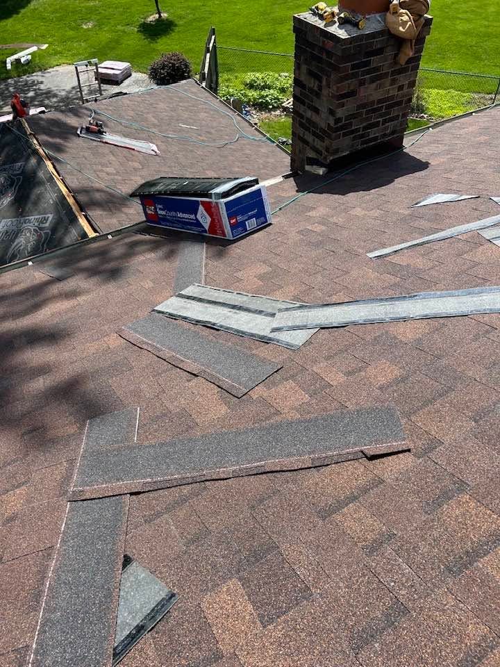 Rooftop with scattered shingles and roofing materials beside a brick chimney and grassy yard below