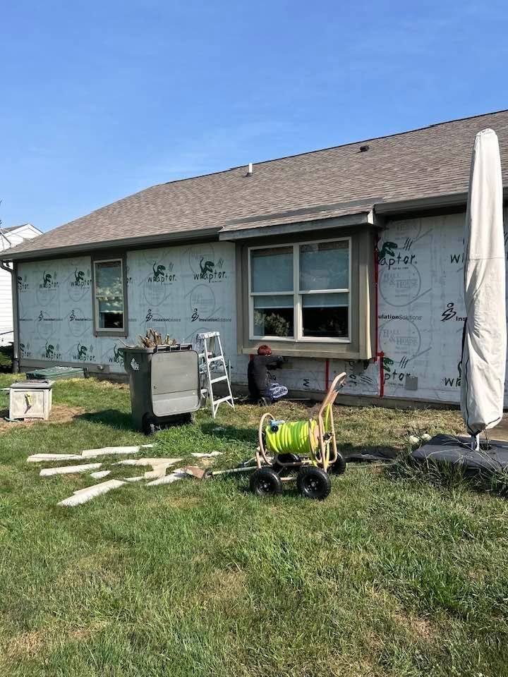 House exterior under construction with wrapped walls, window, lawn tools, and a closed patio umbrella
