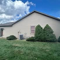 Single-story beige house with a sloped roof, green lawn, shrubs, and a cloudy blue sky.
