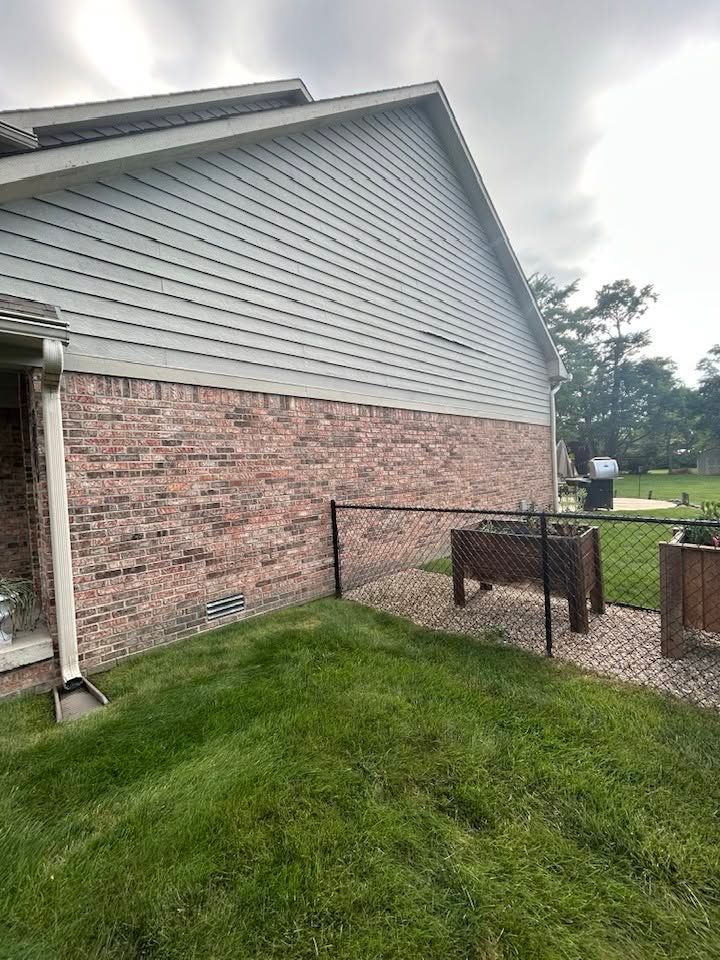 Side of a house with gray siding and brick lower wall beside a fenced grassy yard.
