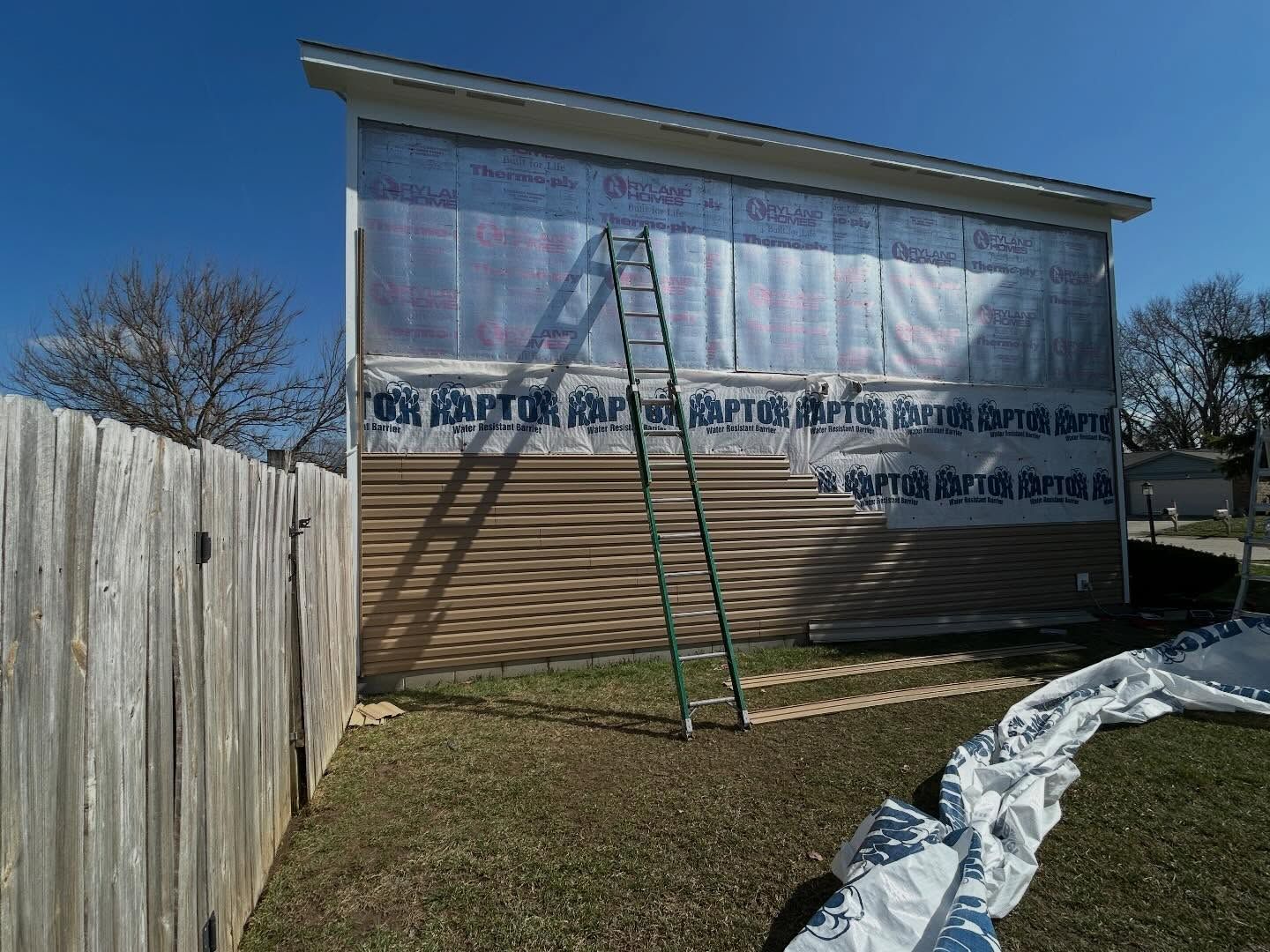 House under renovation with ladder, wood fence, and plastic sheeting on the ground under a blue sky