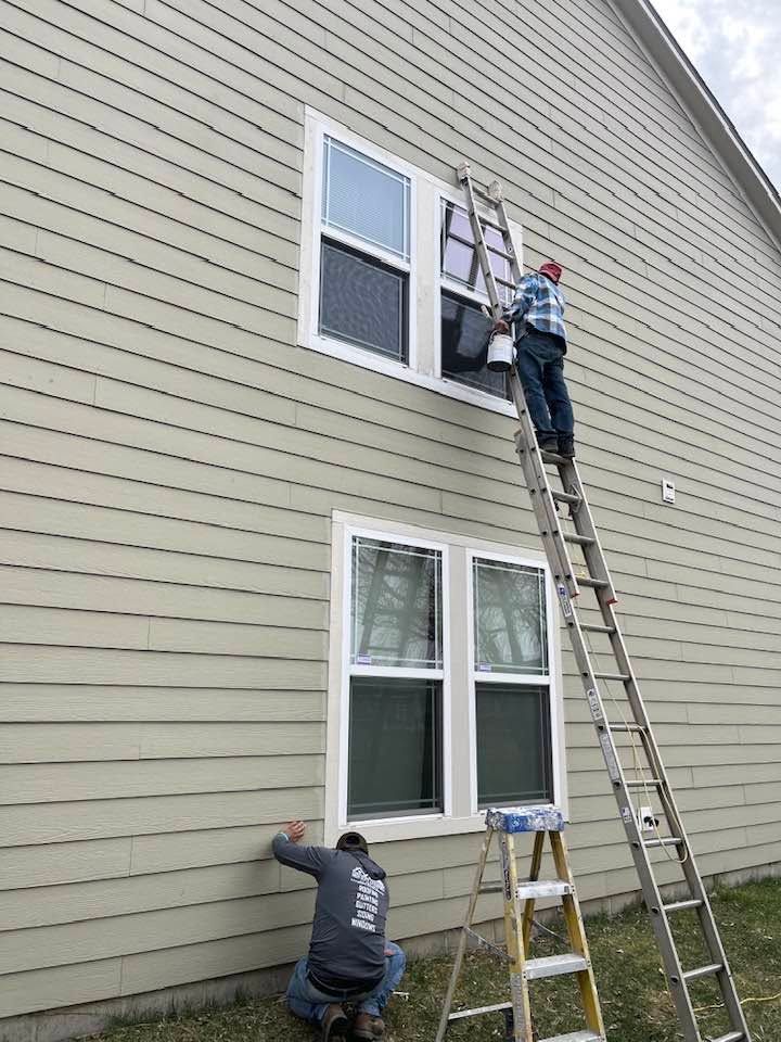 Two workers on ladders installing an upper window on a house exterior