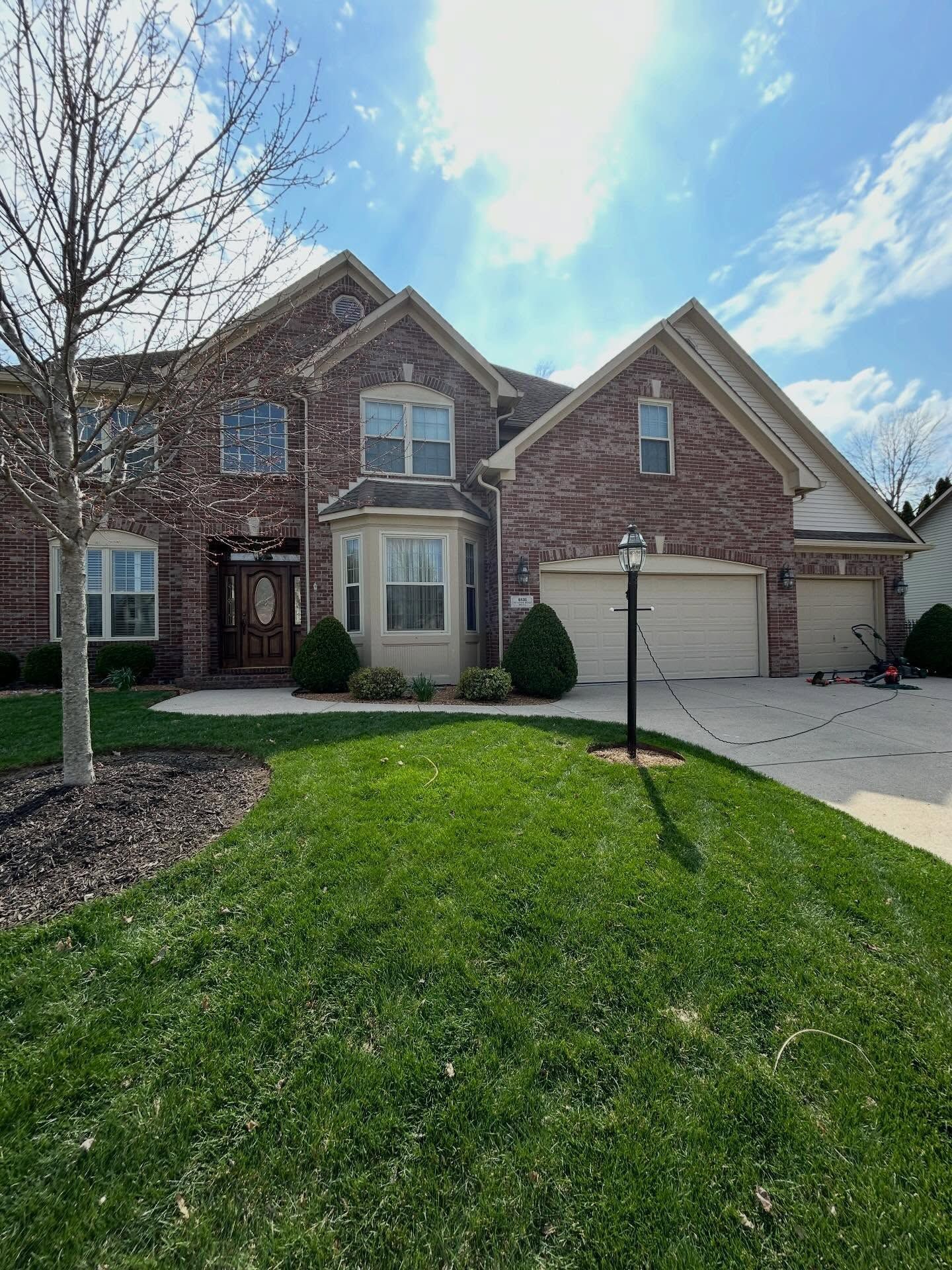 Brick suburban house with two-car garage, front lawn, and leafless tree under a partly cloudy sky
