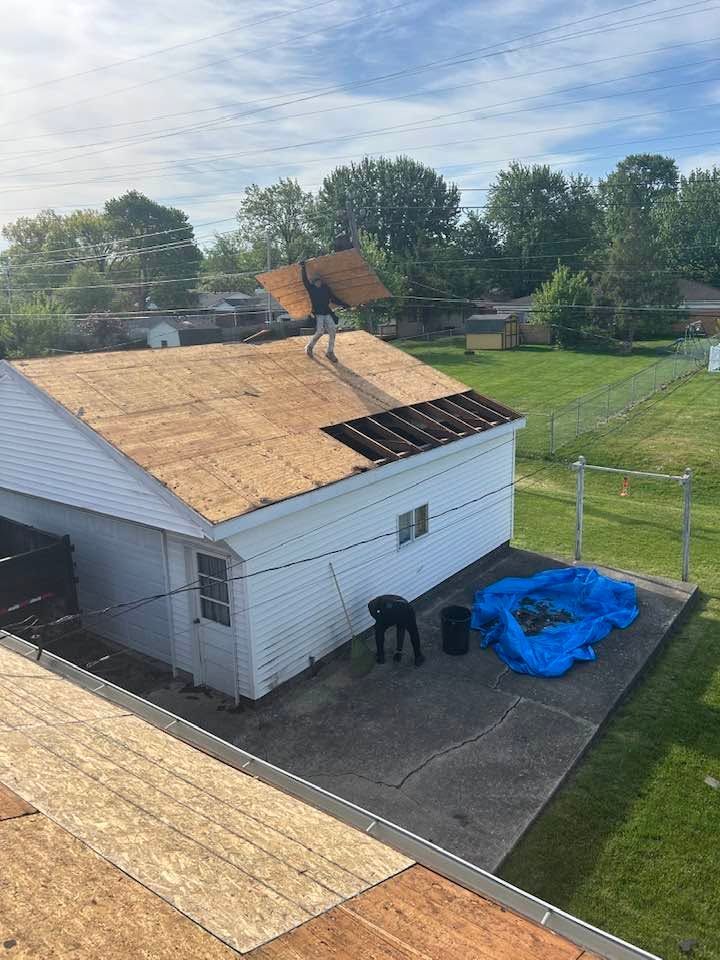 Workers reroofing a white garage with plywood and shingles, blue tarp on the driveway.