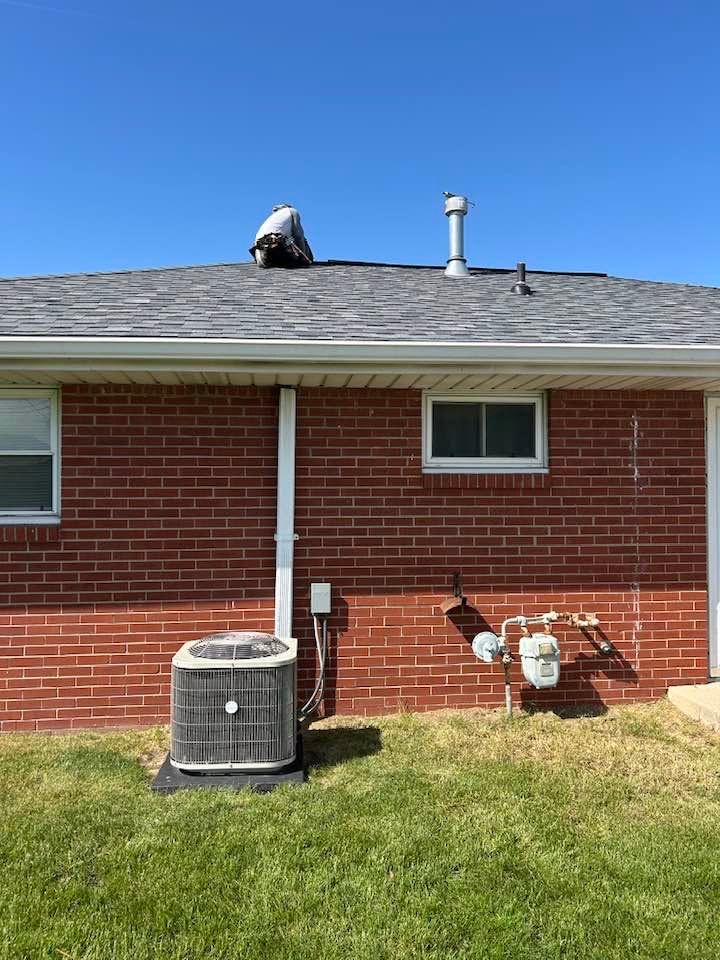 Person crouching on a shingled roof above a brick house with HVAC units in the yard