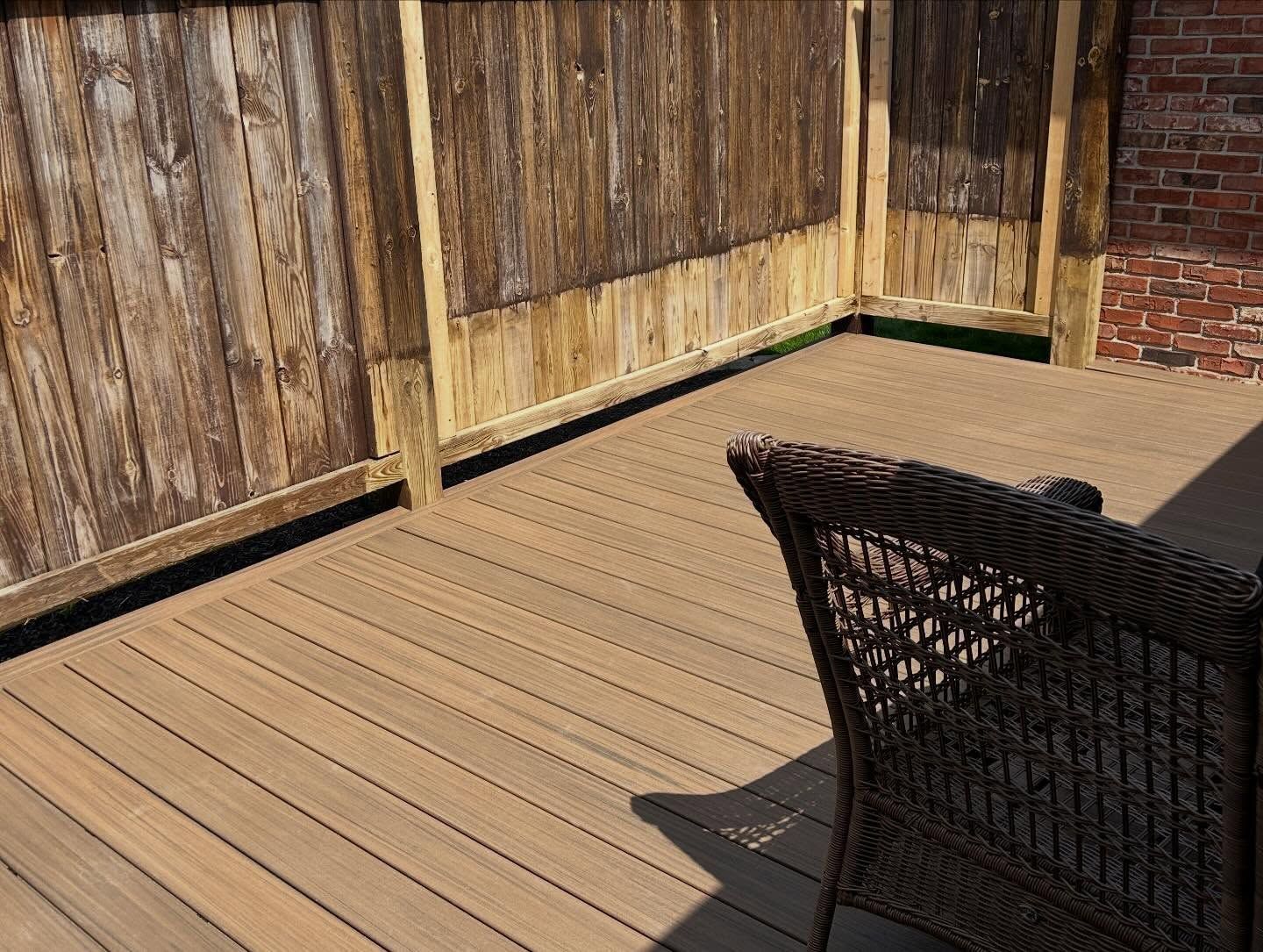 Wooden patio deck beside a weathered fence, with a wicker chair in the foreground.