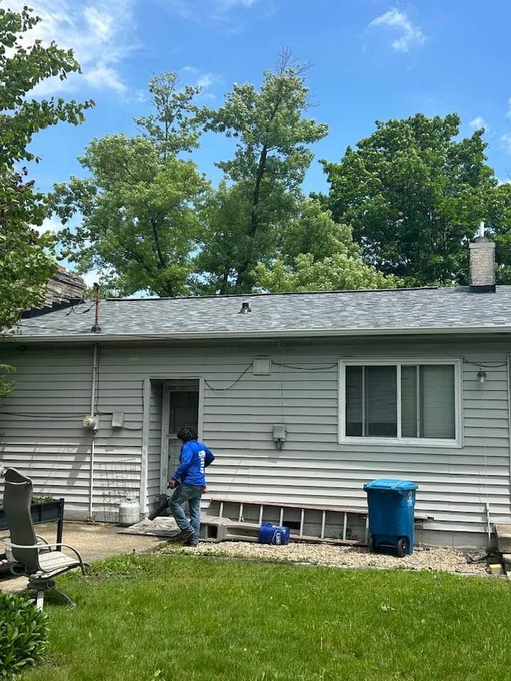 Person in blue shirt working outside a gray house with a green lawn and trees under a blue sky