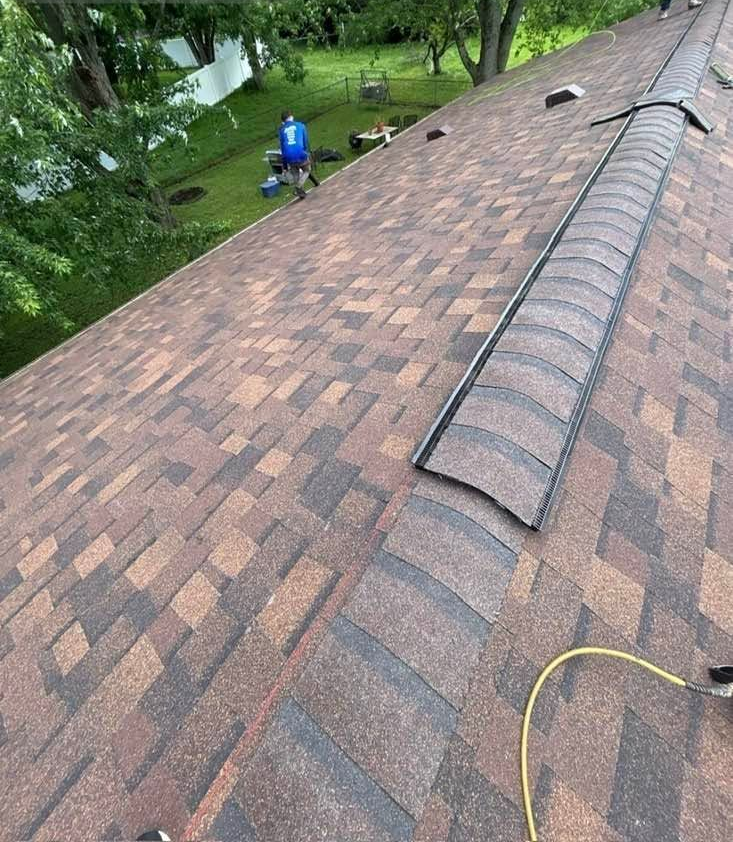 Roof shingles with a ridge vent and a worker in blue on the ground below, surrounded by trees