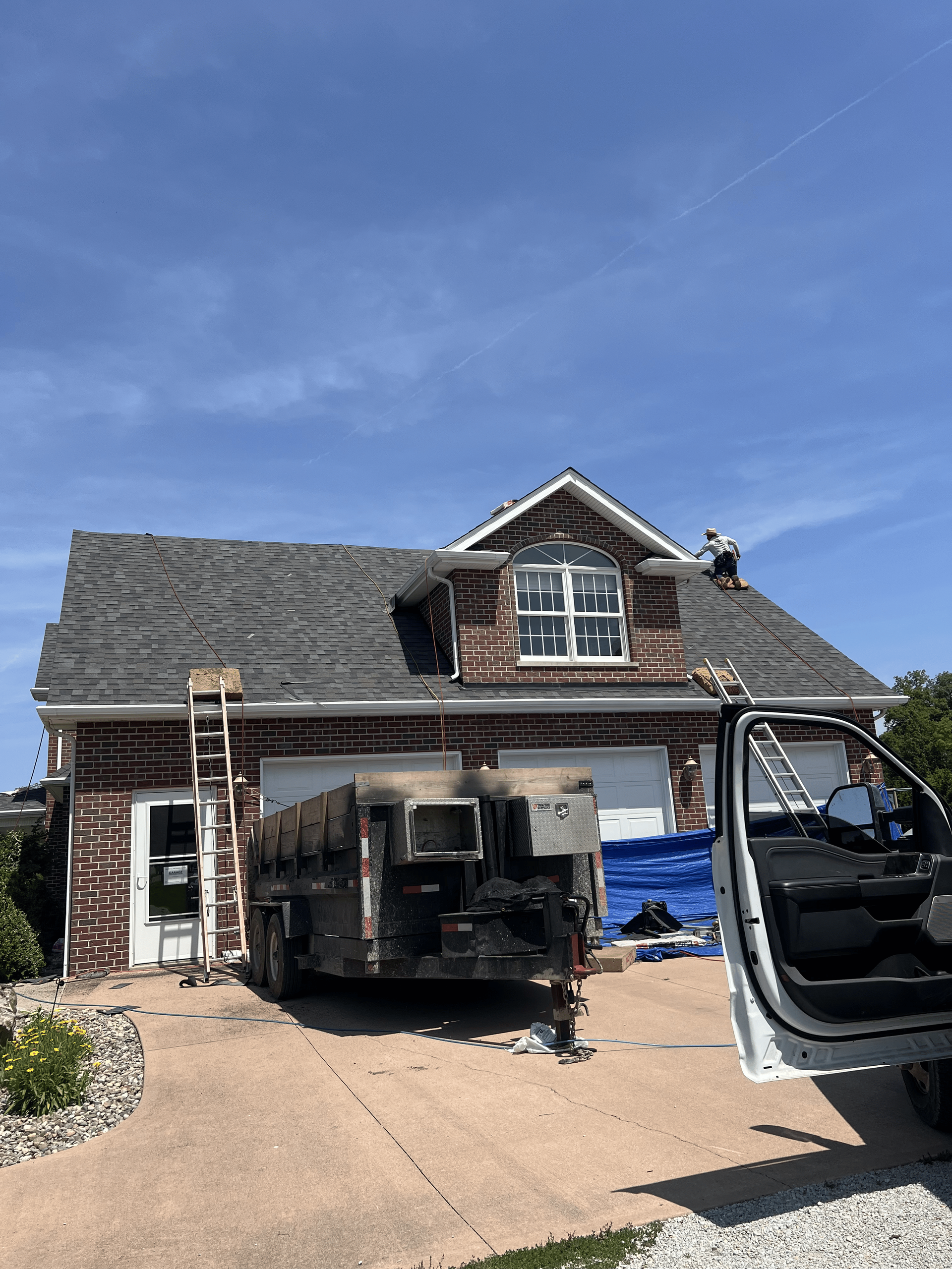 Damaged house with collapsed roof, construction truck in driveway, and open vehicle door under a blue sky