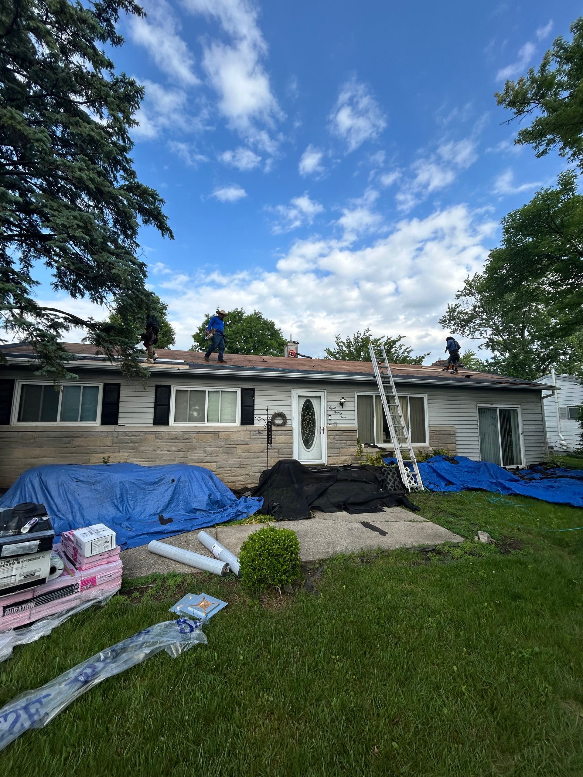 Single-story house under roof repair, with tarps, ladders, and workers on the roof and lawn.