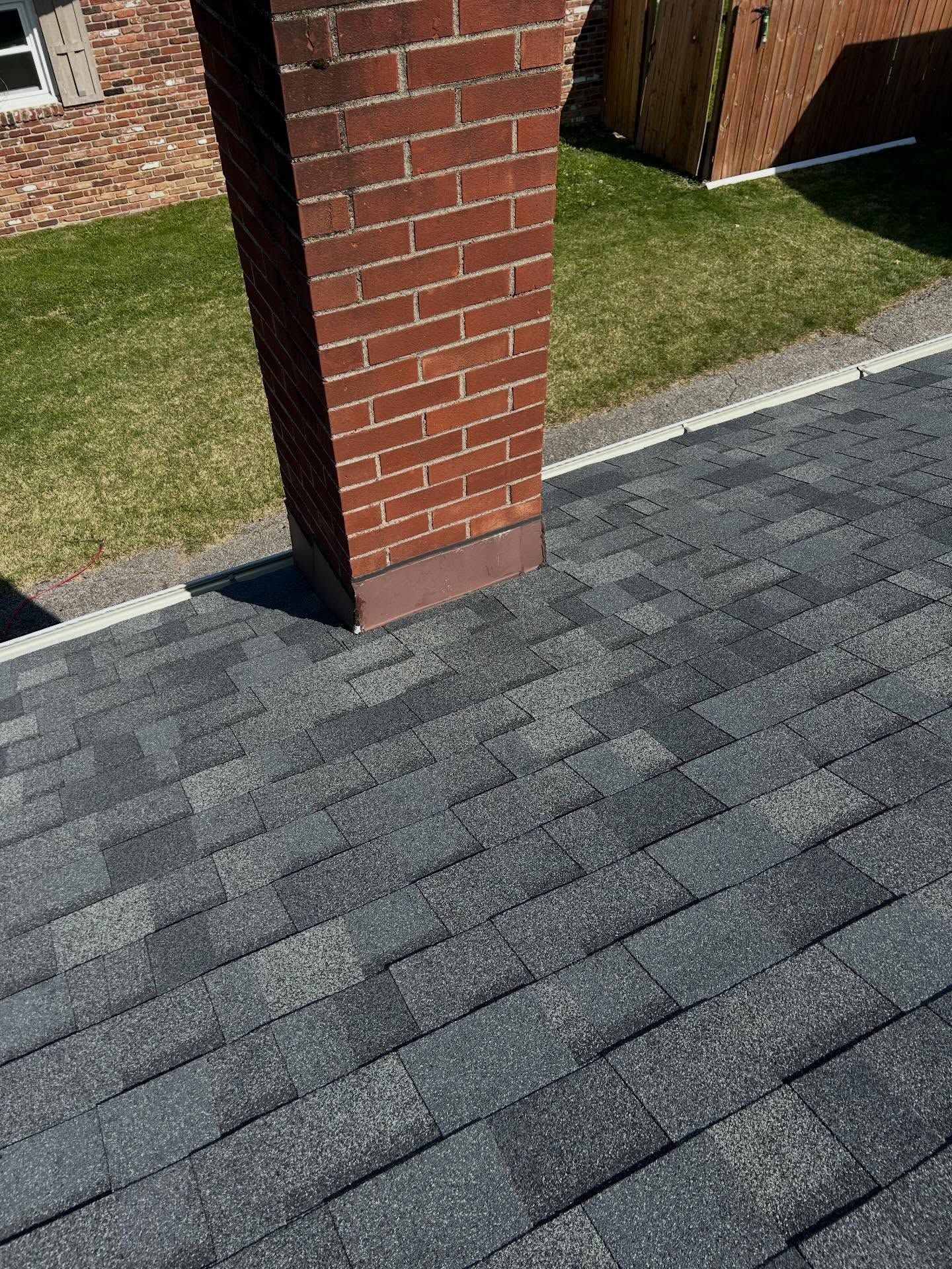 Gray roof shingles beside a brick chimney, with grass and a paved edge in the background