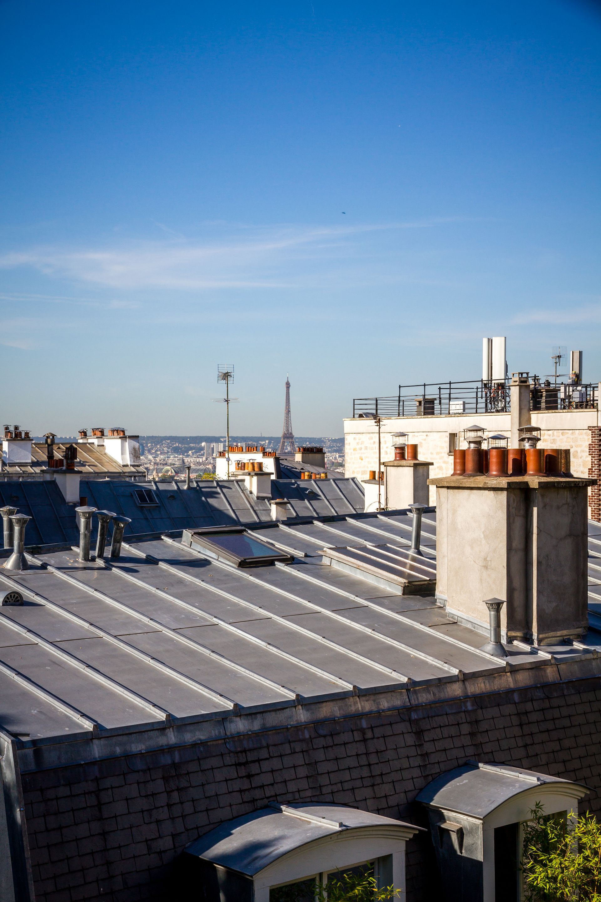 Rooftop with metal chimneys and vents under a clear blue sky, overlooking distant city buildings