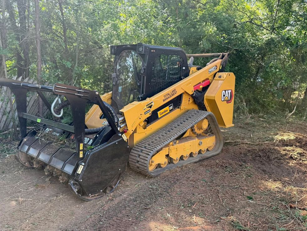 Yellow skid steer with brush mower attachment working in a wooded area.