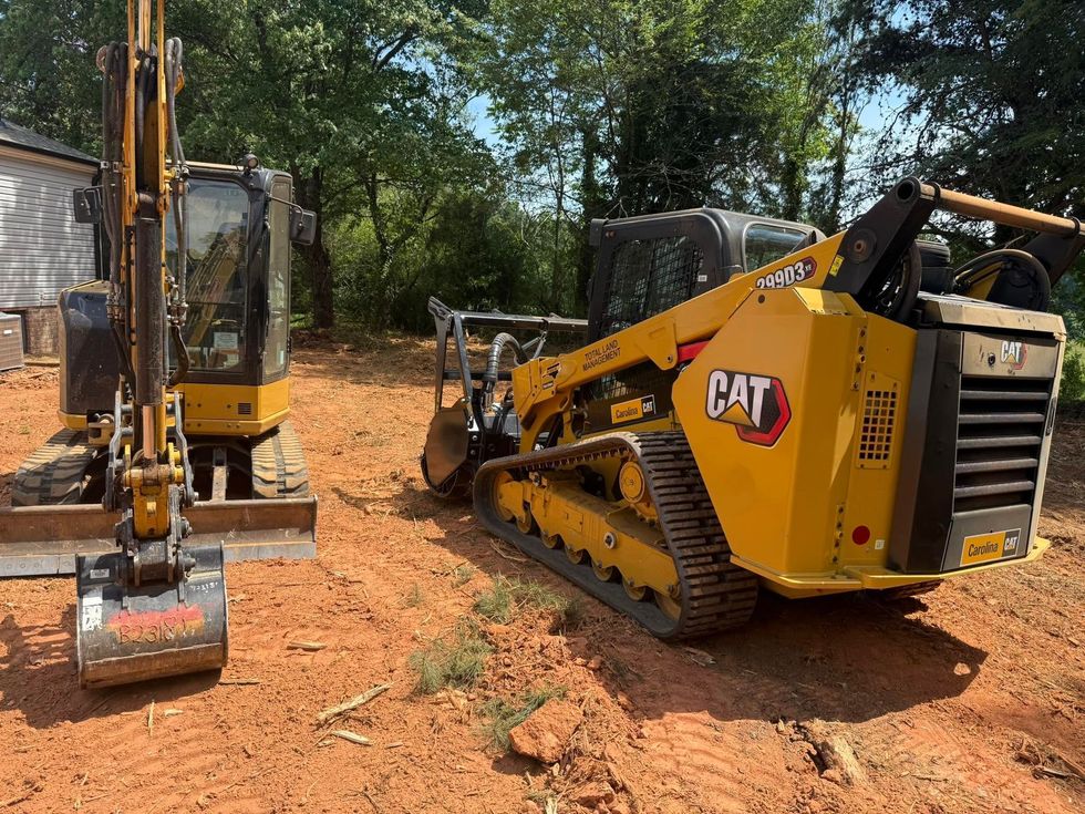 Yellow CAT skid-steer and excavator parked on a dirt clearing under trees.