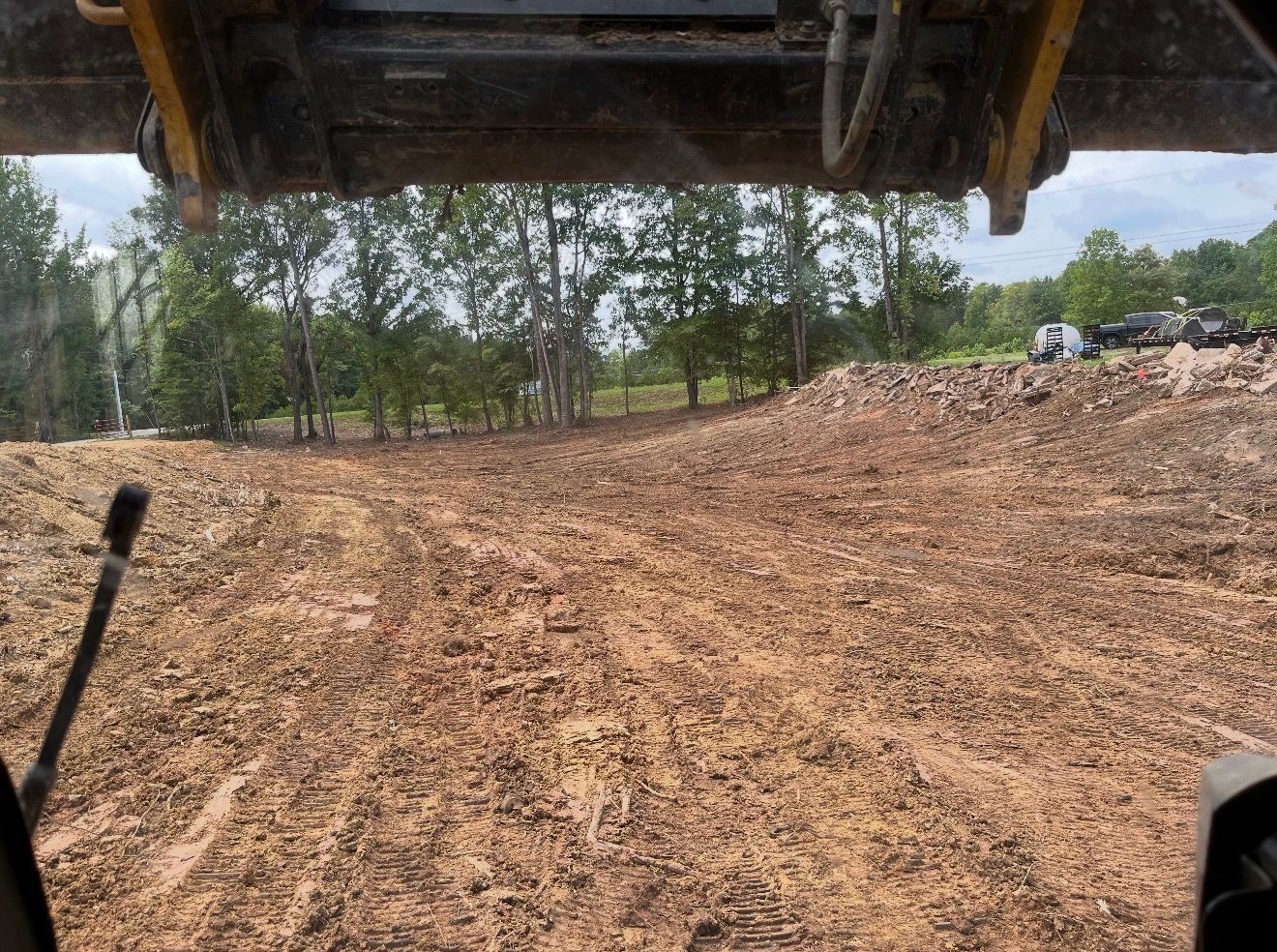 Dirt-covered construction site seen from a heavy machine, with cleared ground and trees in the background.