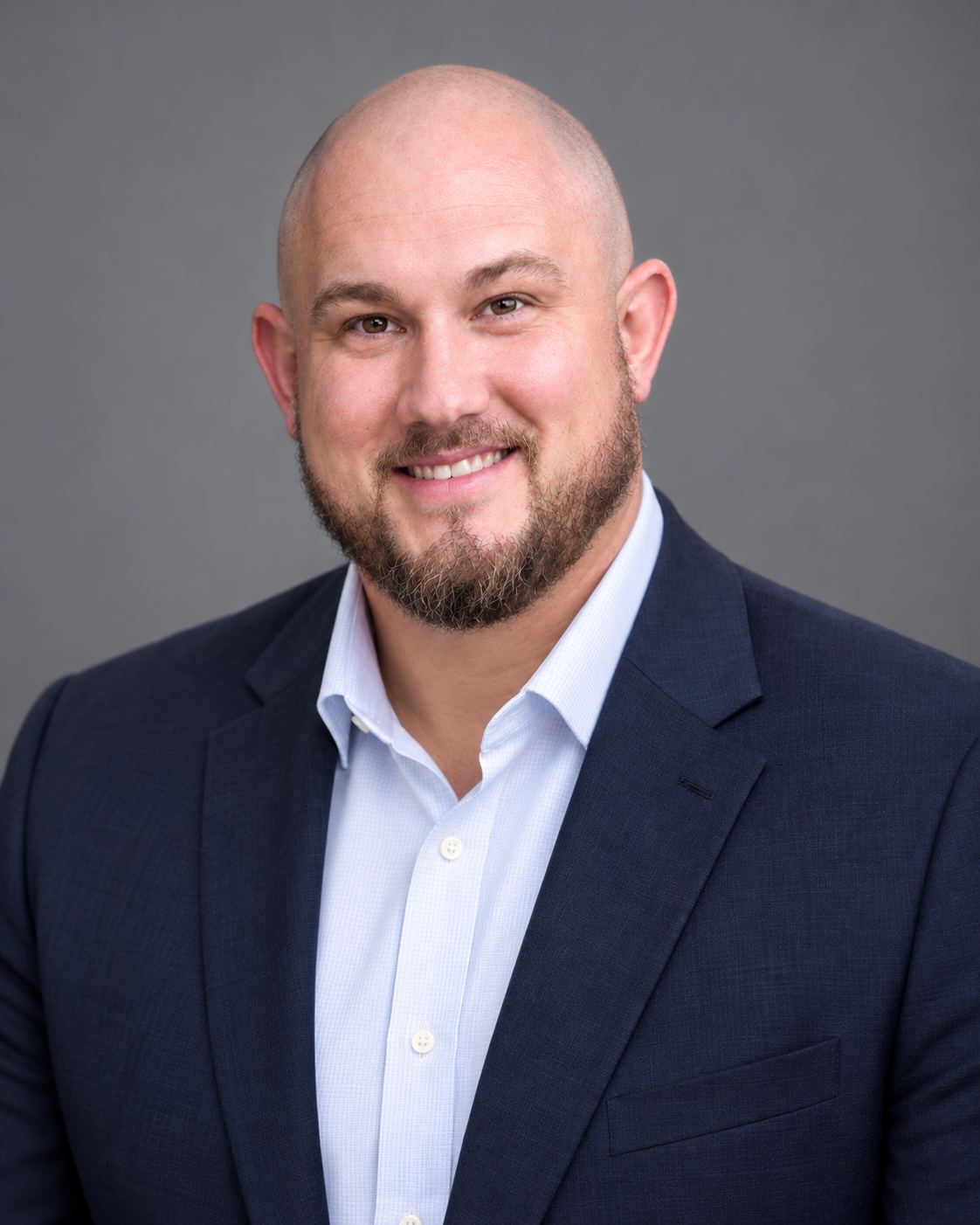 Smiling bald man in a navy blazer and light blue shirt against a gray studio backdrop