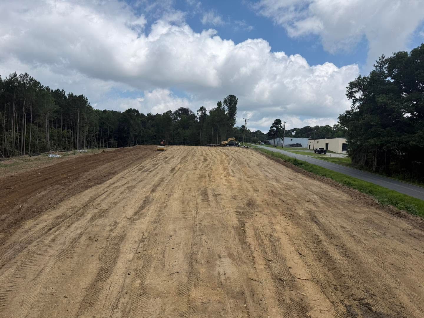Cleared dirt road under cloudy sky, with trees lining the sides and buildings in the distance