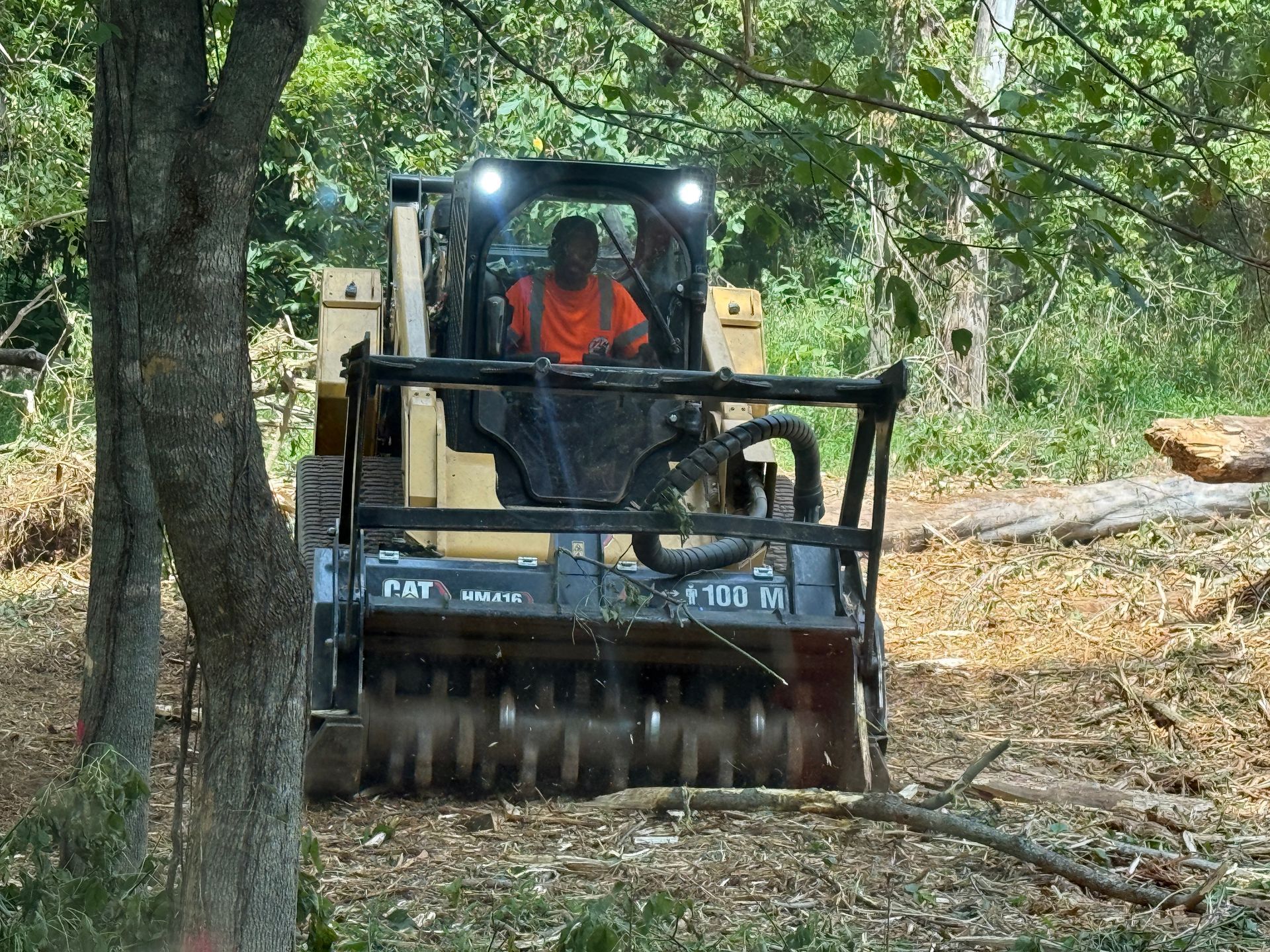 Compact track loader clearing brush in a wooded area, with an operator in an orange shirt.