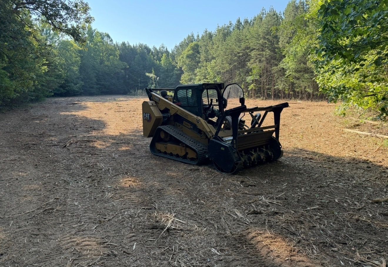 Tracked skid steer on a wooded dirt clearing, with a person operating it on the right side.