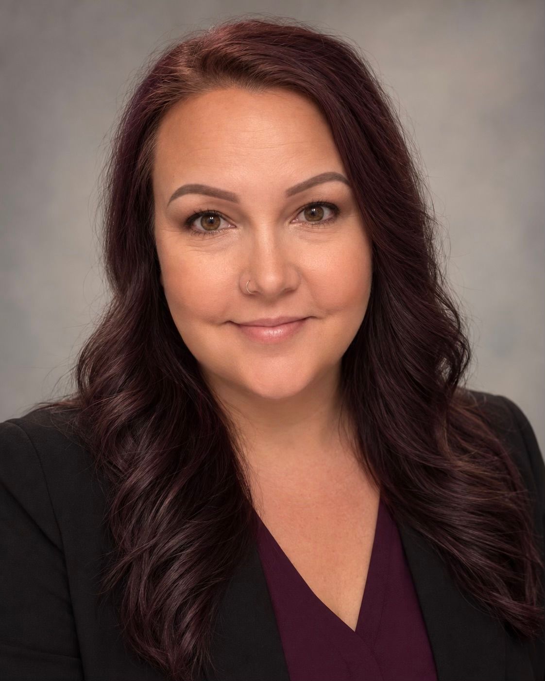 Studio headshot of a woman with dark wavy hair, wearing a black blazer and plum top, smiling softly