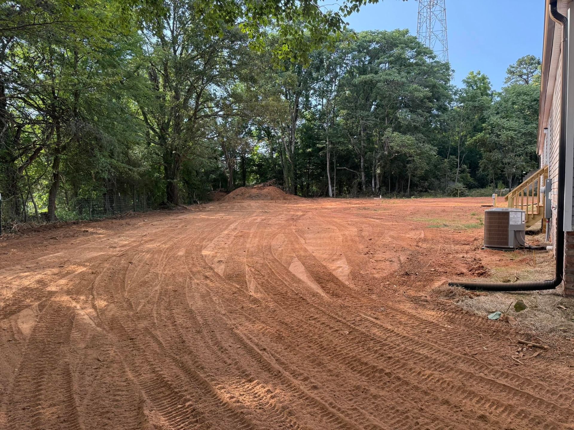 Dirt clearing beside a brick building, with trees lining the background and an air conditioner unit on the right.