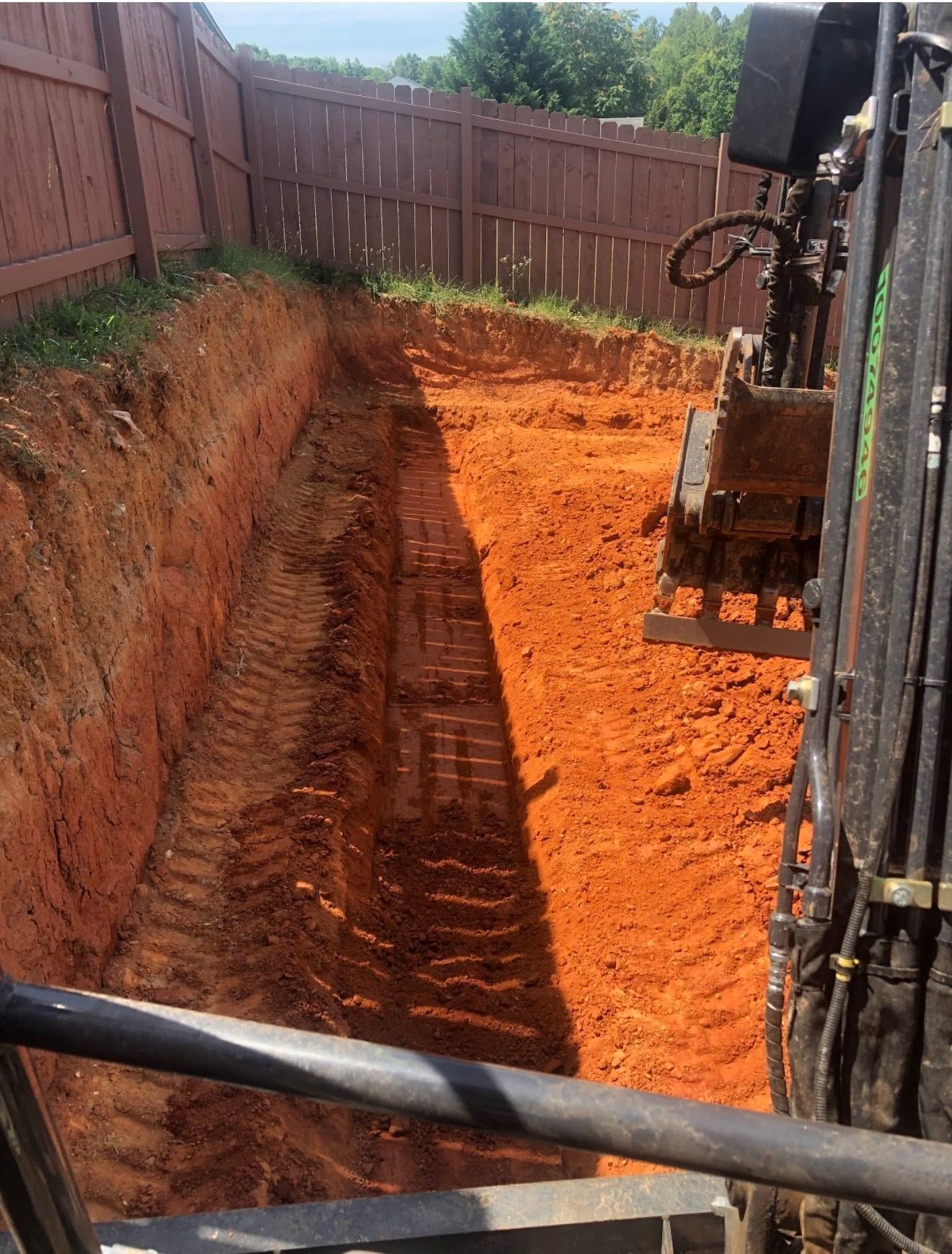 Backyard excavation site with a deep trench, orange dirt, and a mini excavator by a fence