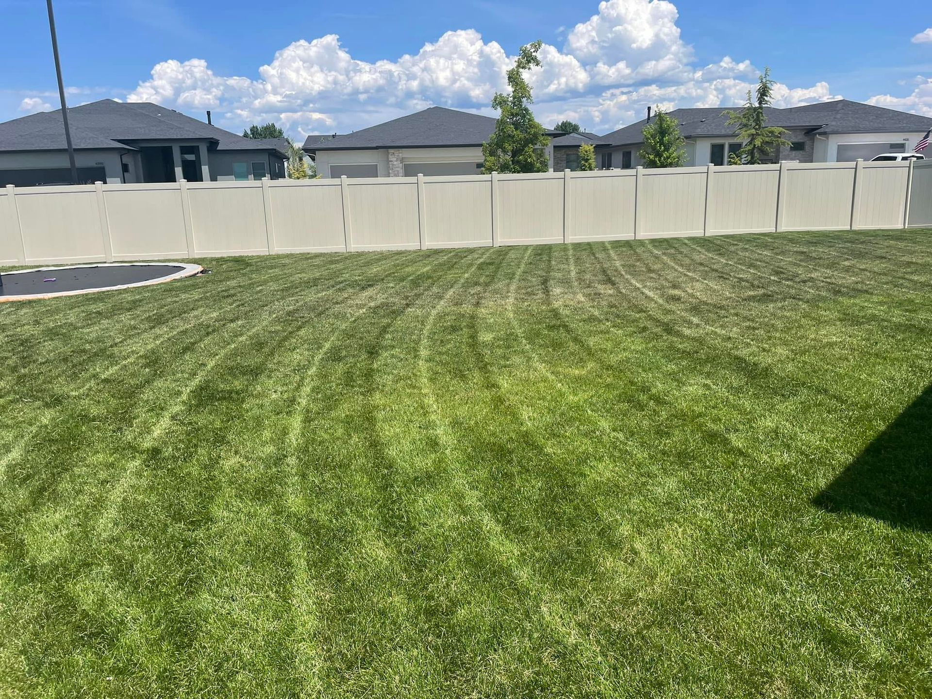 Backyard lawn with a white fence, suburban houses, and a partly cloudy sky