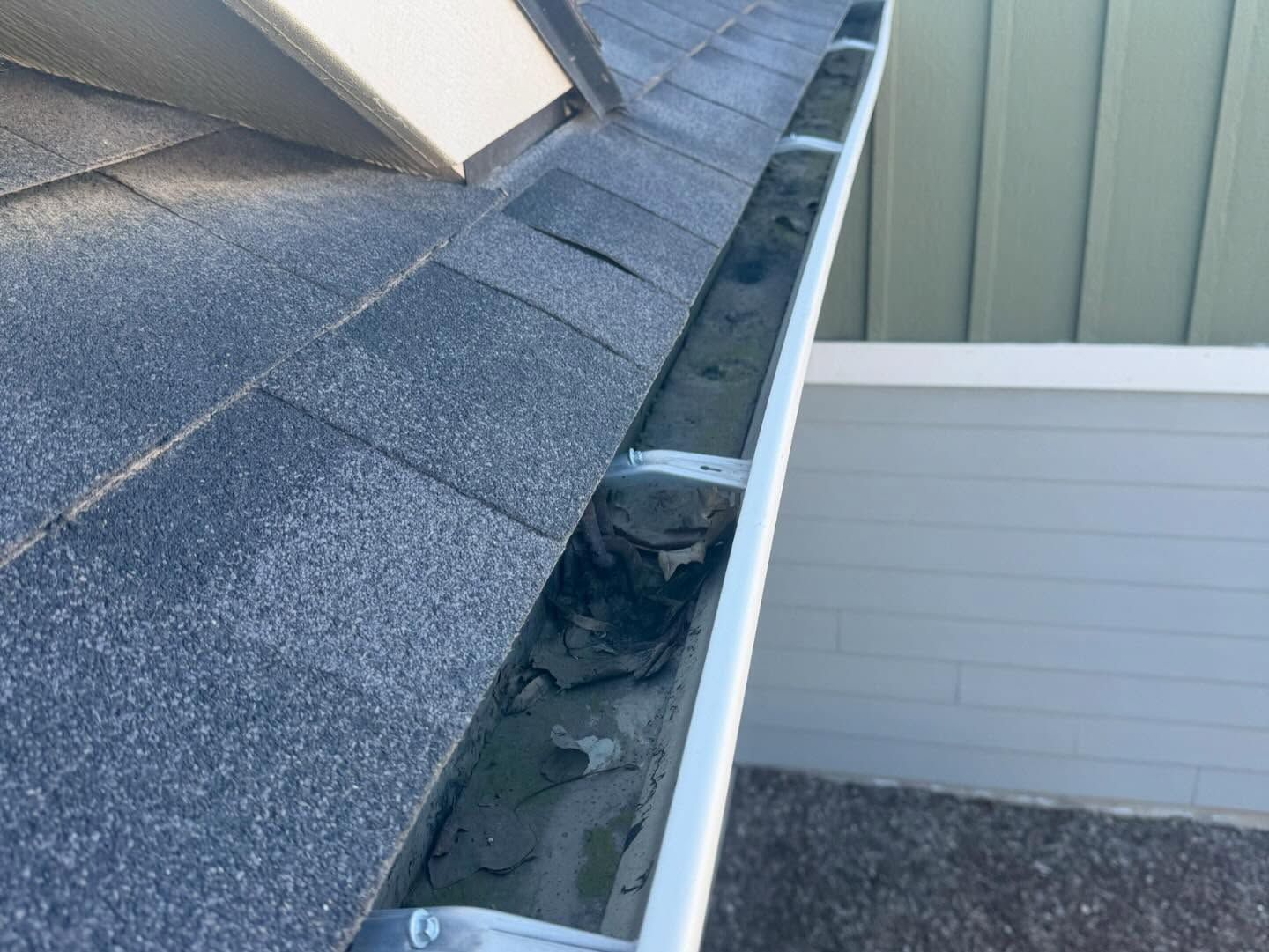 Damaged roof gutter with debris beside gray shingles and white siding