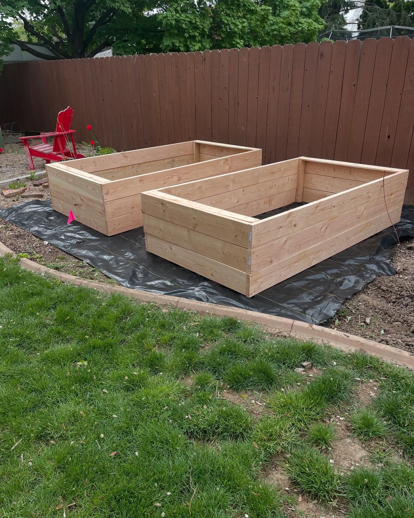 Two unfinished wooden raised garden beds beside a brown fence on black landscaping fabric.