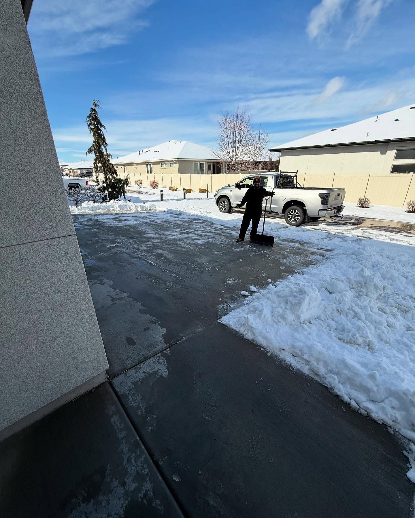 Person stands on snowy pavement beside a white pickup near beige buildings under a bright blue sky.