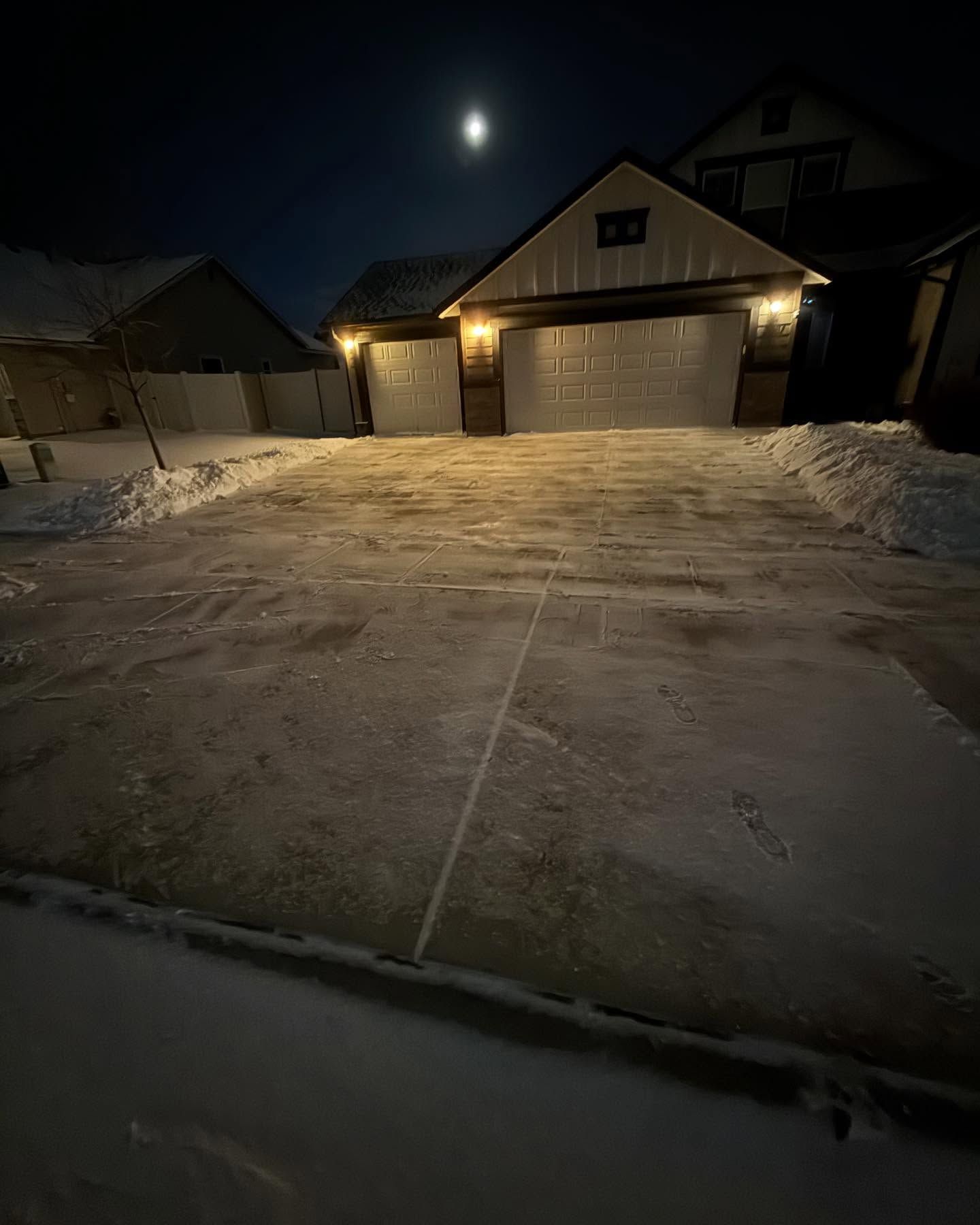 Snow-covered driveway at night leading to a lit garage with a bright moon overhead