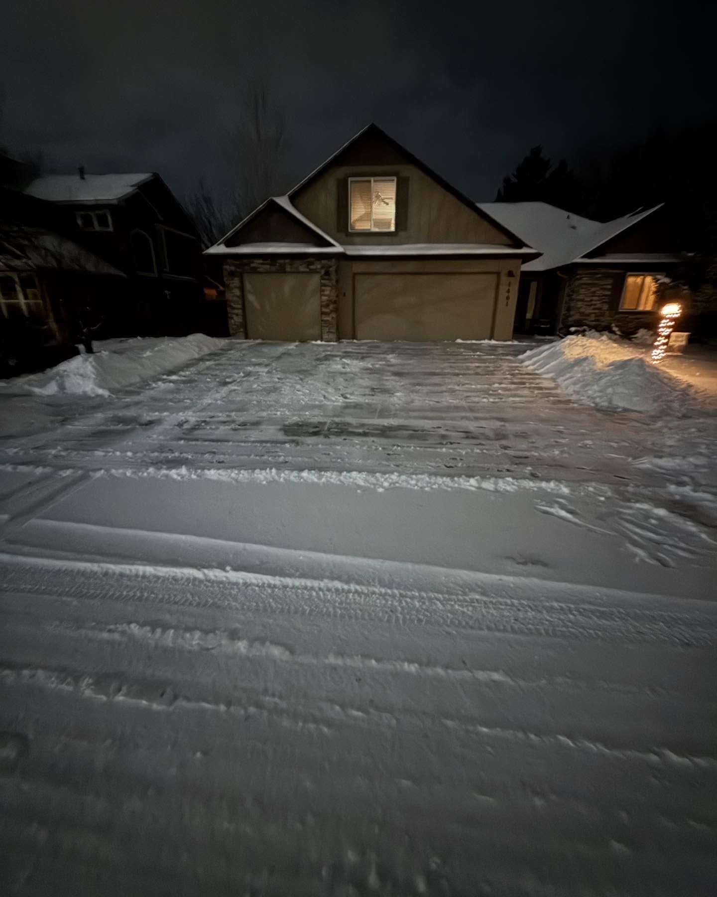 Snow-covered suburban driveway at night with a lit house and garage under a dark sky