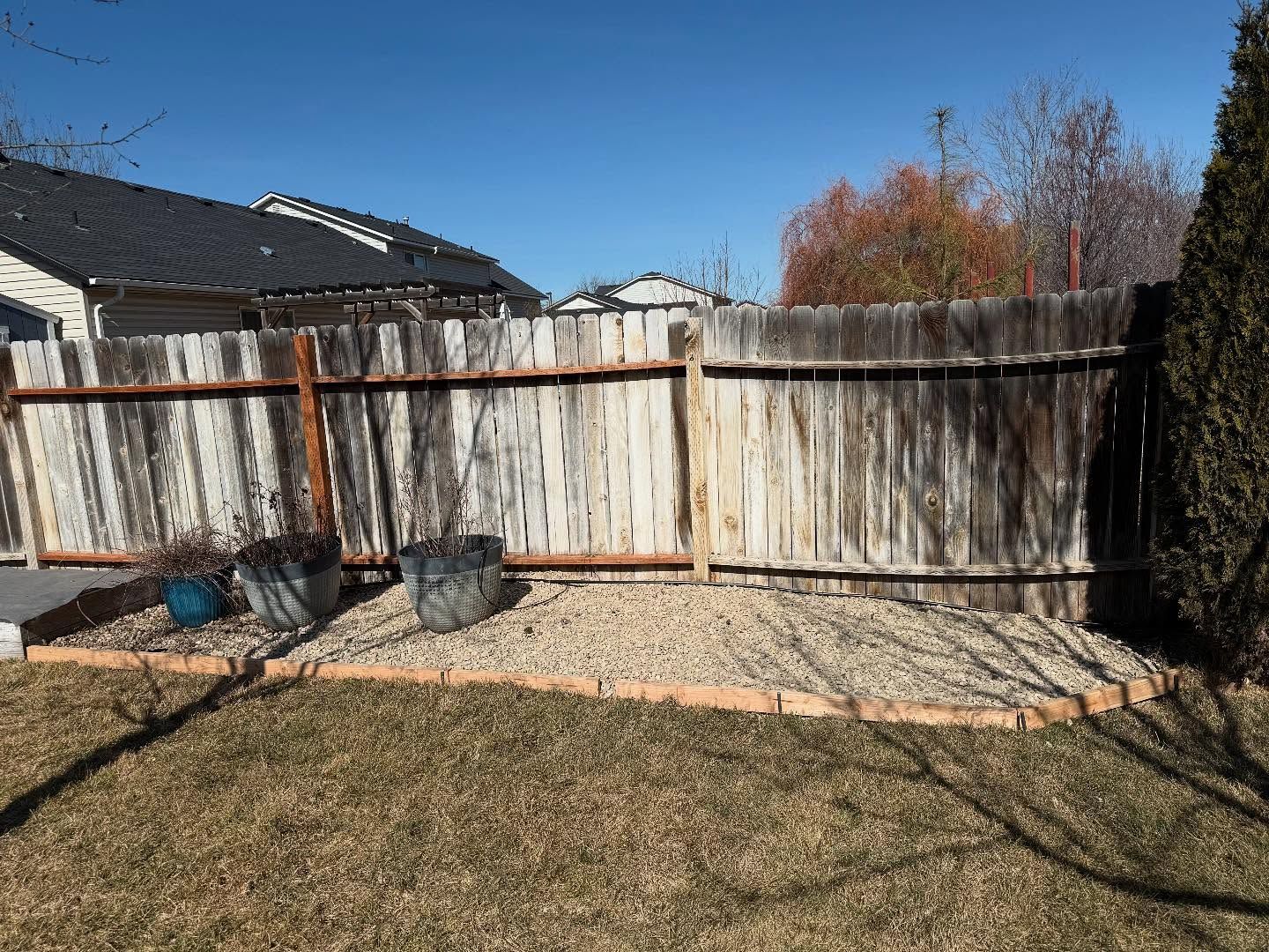 Backyard with a weathered wooden fence, gravel strip, and dry grass under a clear blue sky.