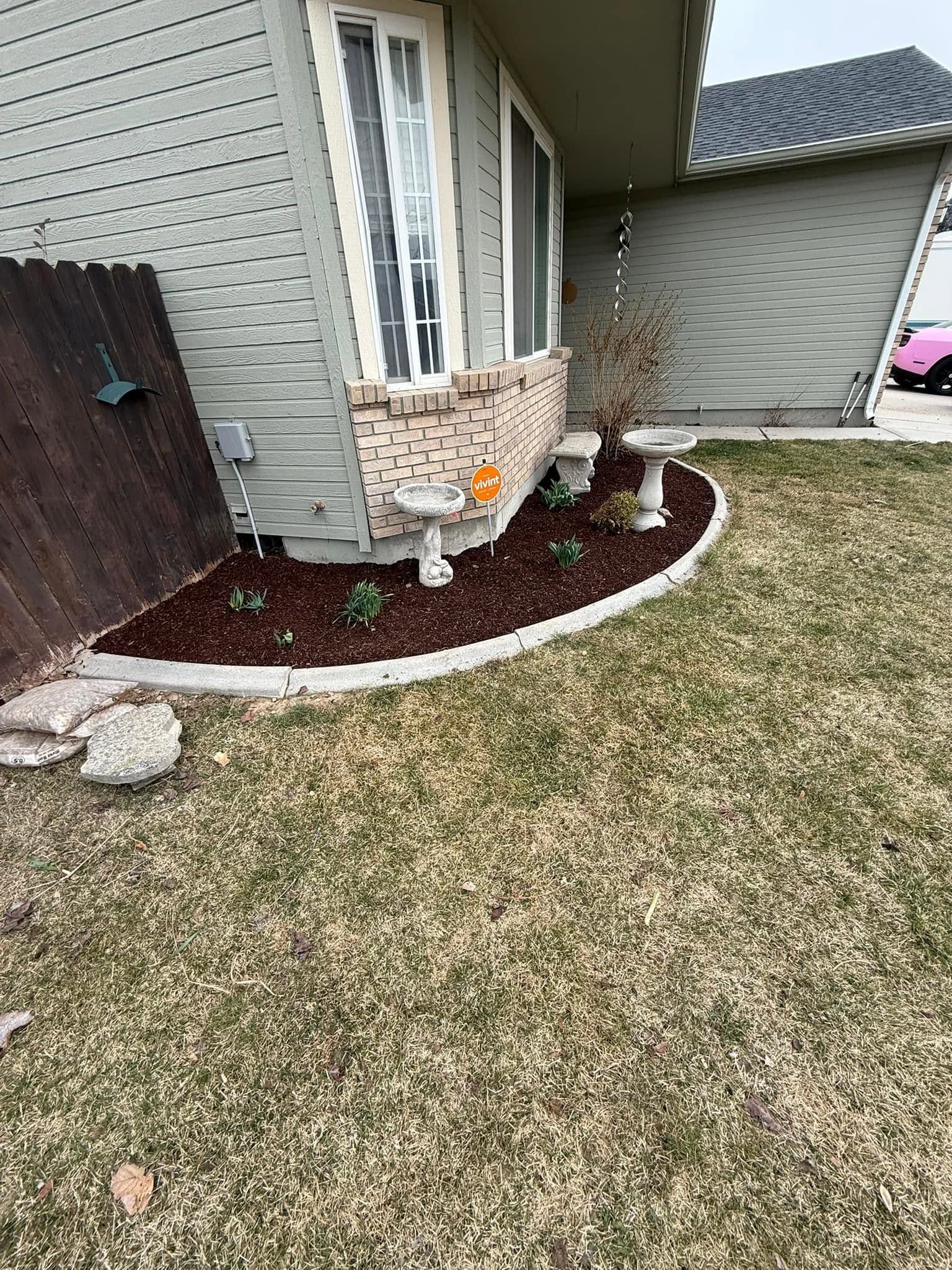 House corner with curved mulched flower bed, small plants, and a brick planter beside a lawn