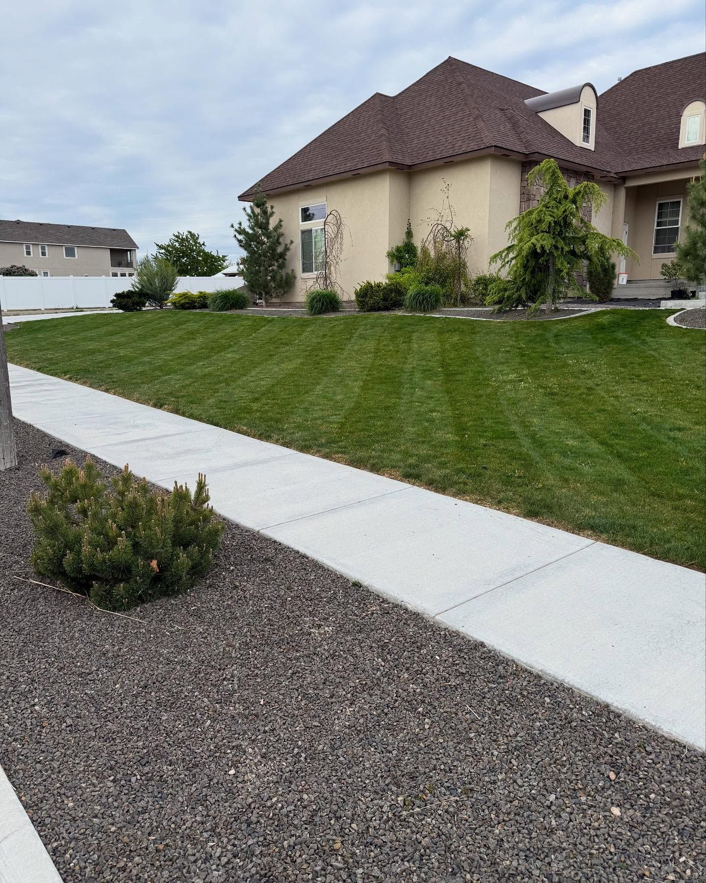 Suburban house with brown roof, green lawn, concrete sidewalk, and gravel landscaping under cloudy skies
