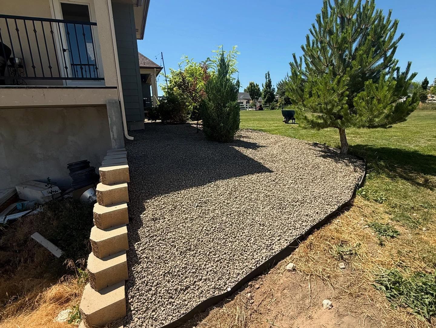 Gravel landscaping bed beside a house, bordered by a curved black edge and young trees under a blue sky