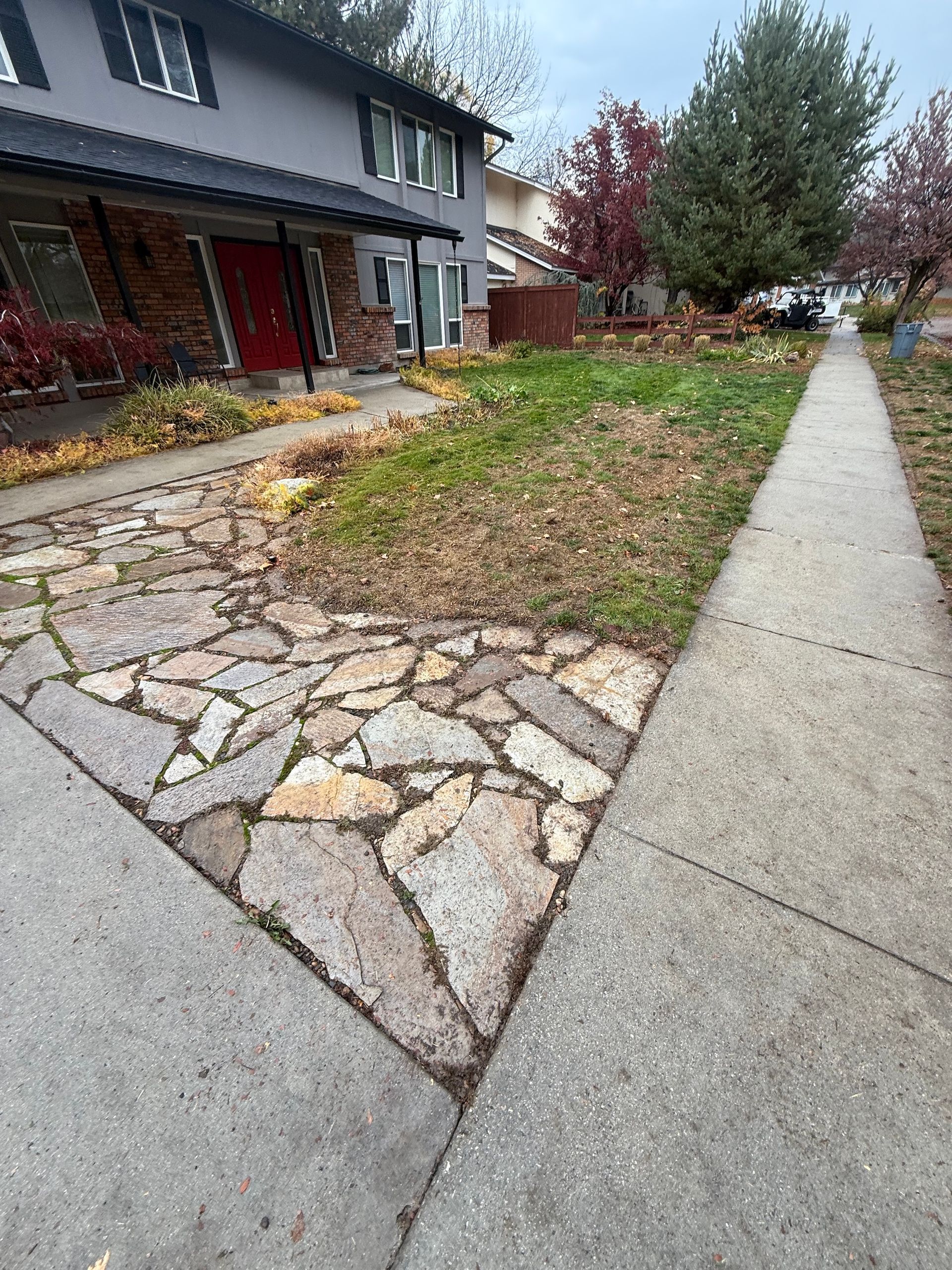 Sidewalk beside a house with a stone-and-gravel yard and autumn trees in the background