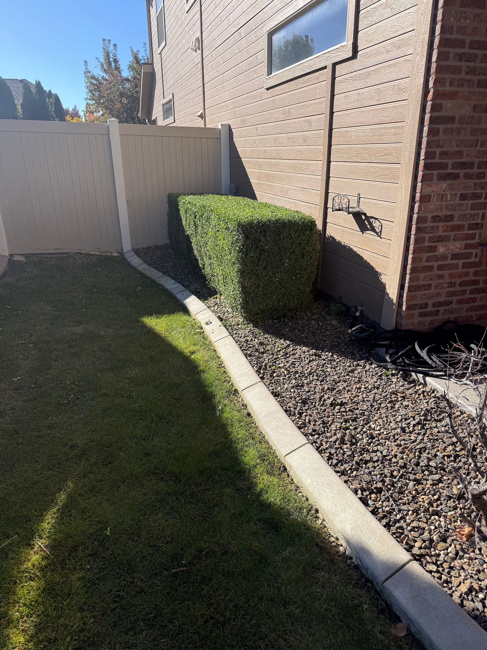 Side yard with brick house, white fence, green lawn, trimmed hedge, and gravel border in sunlight.