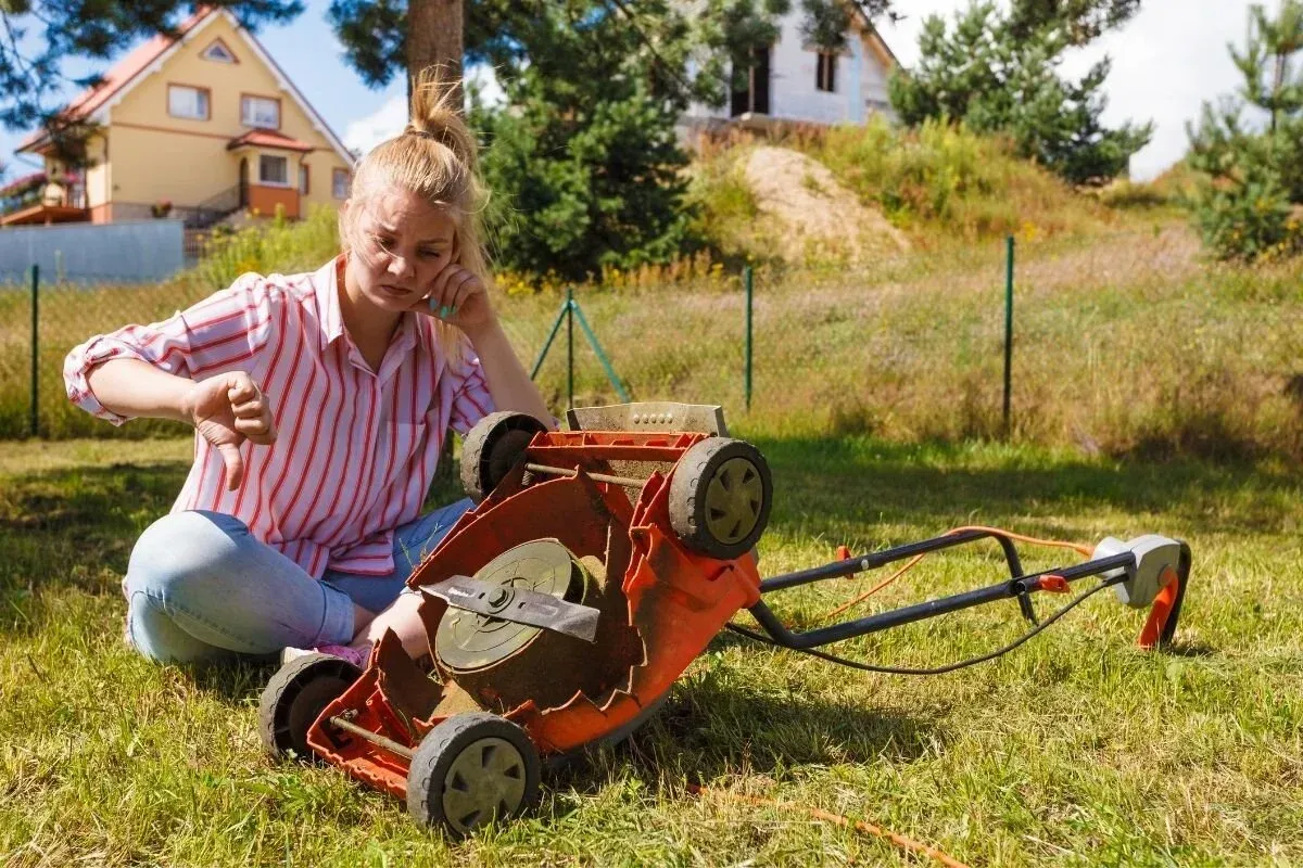 Person kneeling beside an orange lawnmower in a grassy yard, with a house in the background