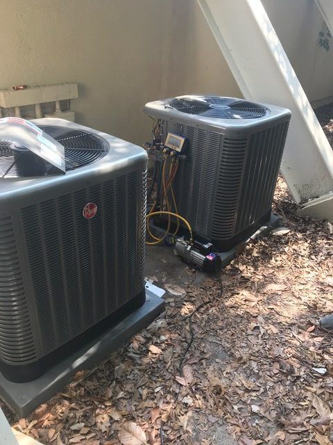 Two air conditioning units on gray pads, outdoors near foliage.