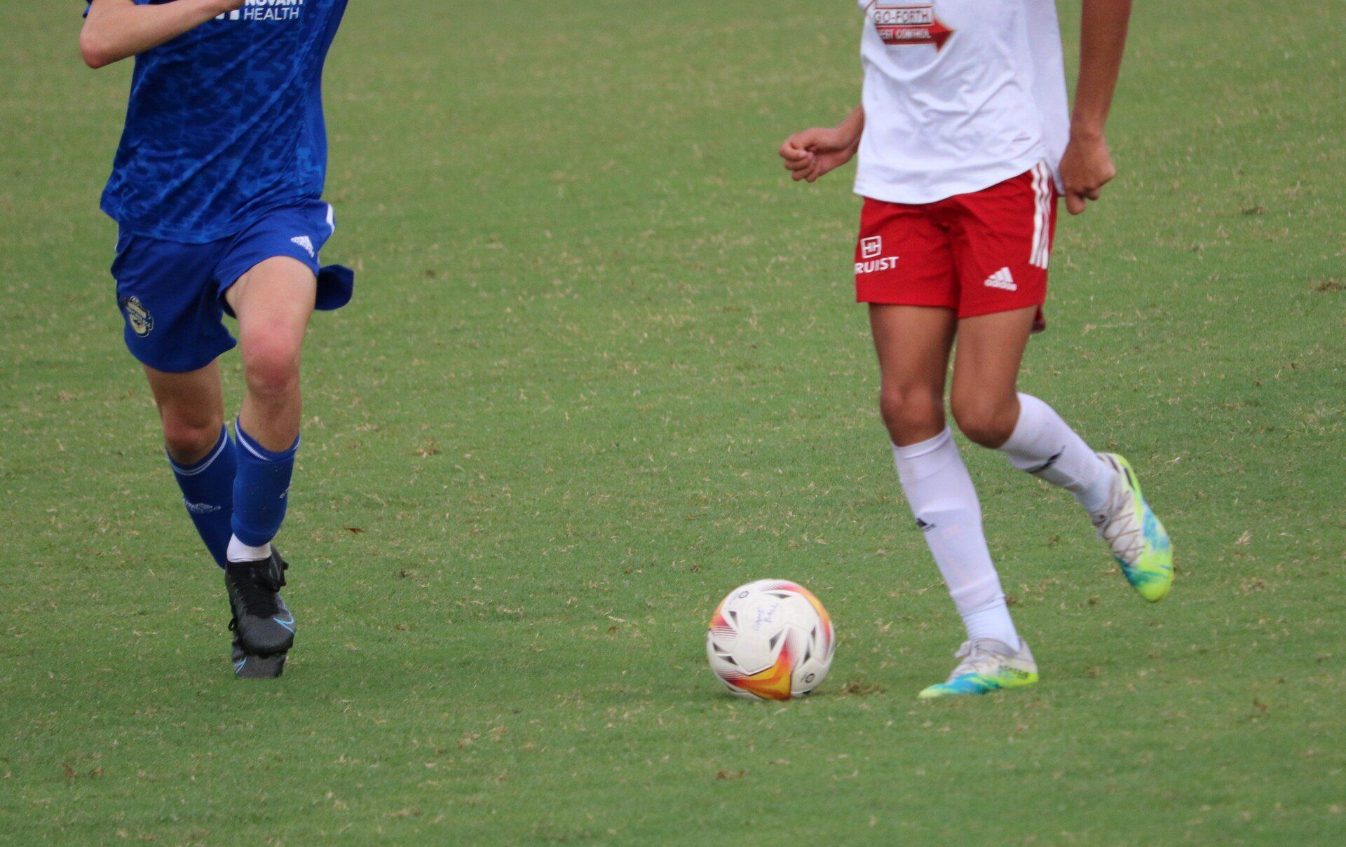 Two soccer players chase a ball on a grass field, one in blue and one in red and white.