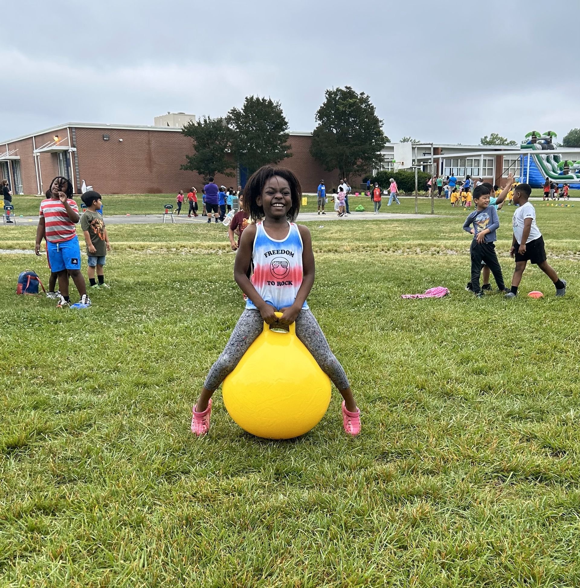 Child smiling on a yellow hopper ball in a grassy school field, with others playing in the background