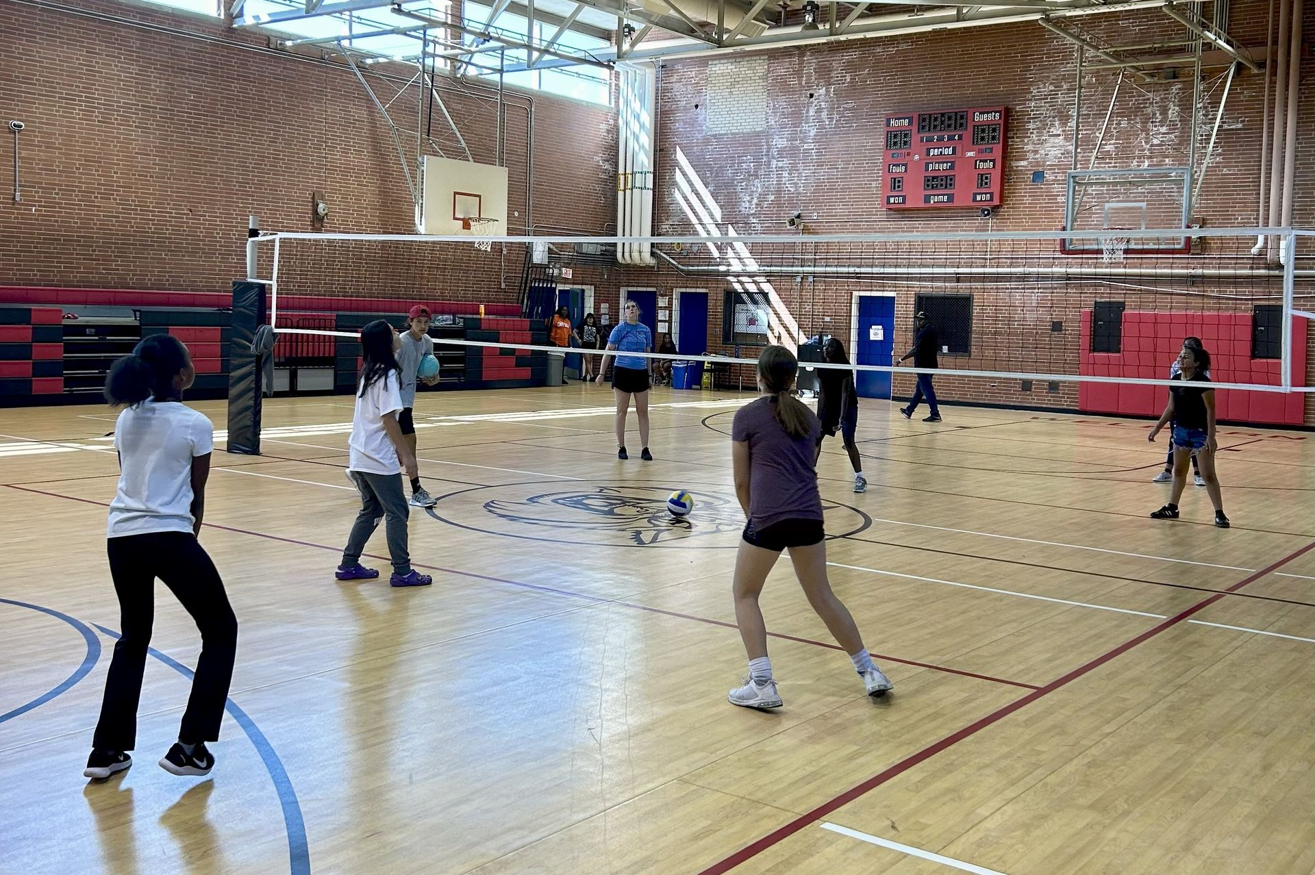 People playing volleyball in a gym with a net, wood floor, and brick walls.