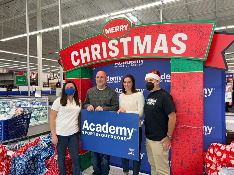 Four people posing under a red Merry Christmas sign at Academy Sports + Outdoors.