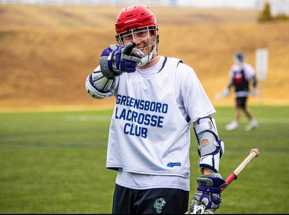 Lacrosse player in red helmet and white Greensboro Lacrosse Club jersey stands on field with stick raised.