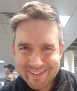 Smiling man in a black jacket indoors, with fluorescent lights and a blurred background.