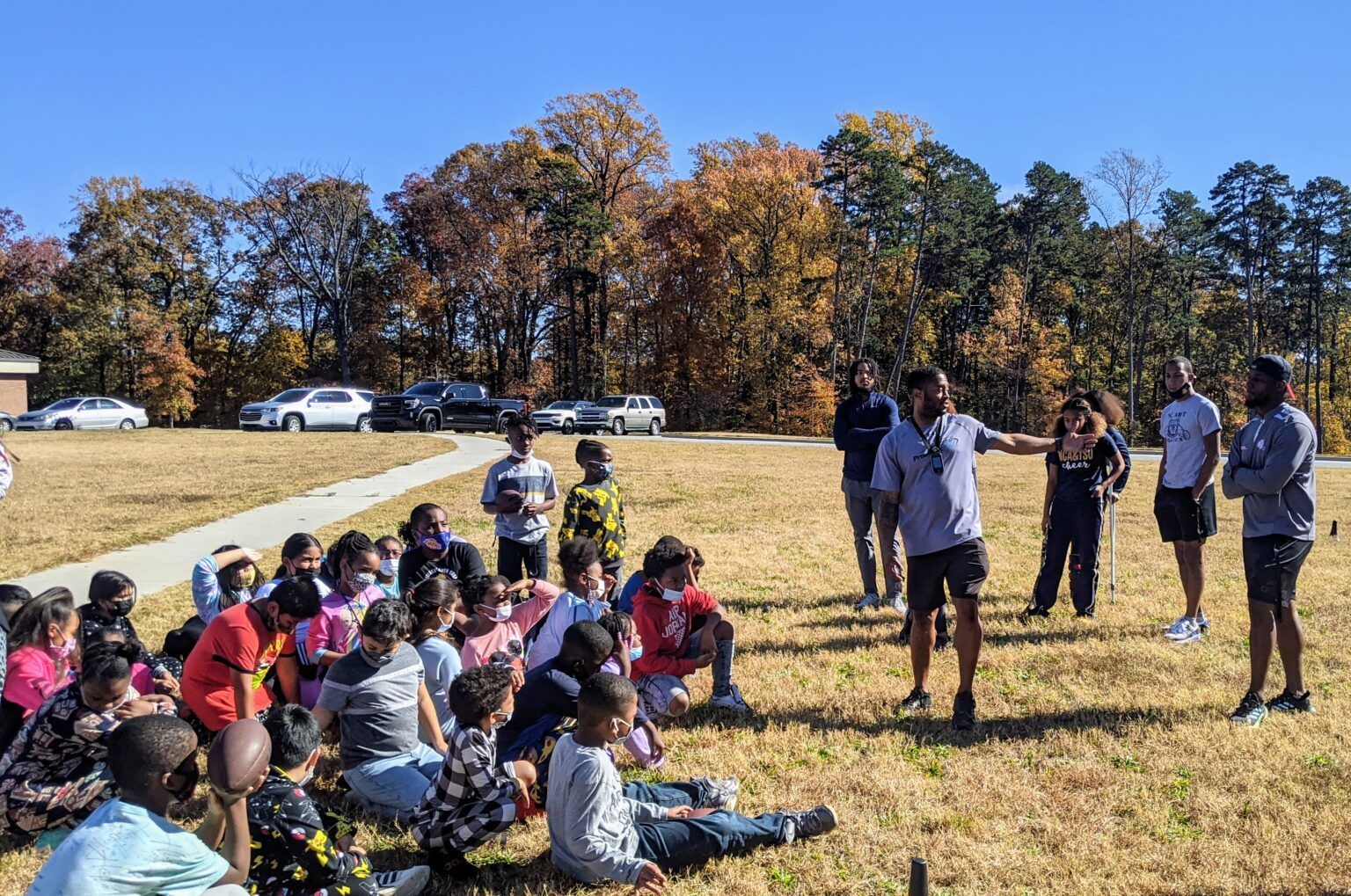 People seated on grass watching a group of speakers outdoors near autumn trees and parked cars