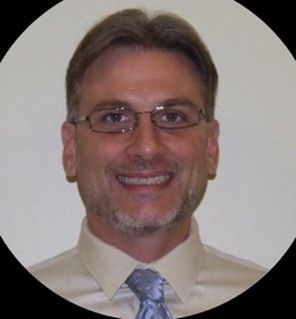 Portrait of a smiling man in glasses, wearing a light shirt and patterned tie on a plain background