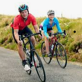 Two cyclists in red and blue jerseys riding on a rural road under cloudy skies