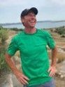 Smiling person in a green shirt and cap standing outdoors near water